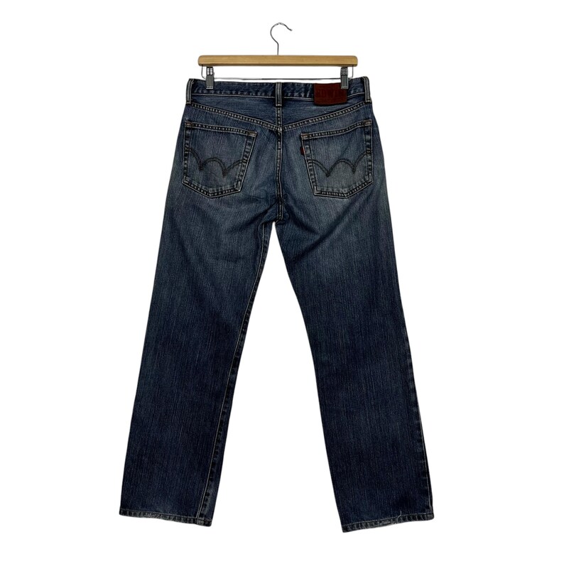 May include: Blue denim jeans with a classic five-pocket design, hanging from a wooden hanger. The jeans have a faded wash, visible stitching, and a leather patch. The back pockets feature a distinctive curved stitch.