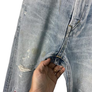 May include: Light blue denim jeans with distressed details, including a large rip and faded patches. The jeans show signs of wear and tear, with visible staining. The stitching is yellow, and the overall look is casual and vintage.