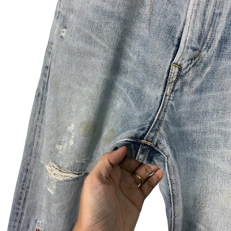 May include: Light blue denim jeans with distressed details, including a large rip and faded patches. The jeans show signs of wear and tear, with visible staining. The stitching is yellow, and the overall look is casual and vintage.