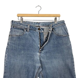 K&ouml;nnte beinhalten: Hellblaue Jeans aus Denim, die an einem Holzkleiderb&uuml;gel h&auml;ngen. Die Jeans haben ein klassisches F&uuml;nf-Taschen-Design, einen Rei&szlig;verschluss und einen Knopfverschluss. Die Jeans bestehen aus strapazierf&auml;higem Denim-Stoff und sind f&uuml;r den Alltag geeignet.