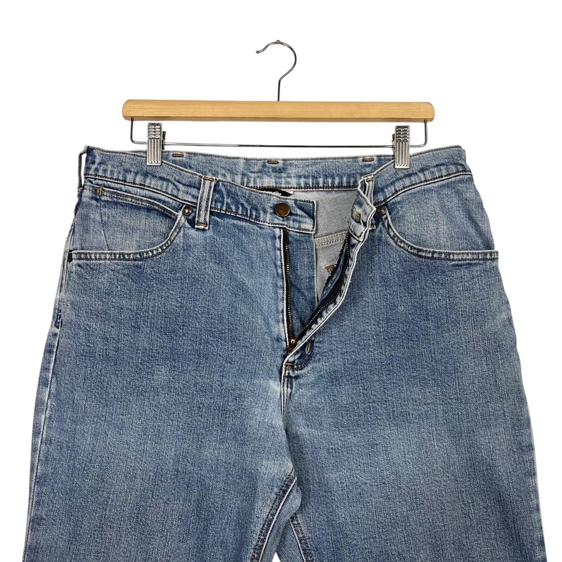 K&ouml;nnte beinhalten: Hellblaue Jeans aus Denim, die an einem Holzkleiderb&uuml;gel h&auml;ngen. Die Jeans haben ein klassisches F&uuml;nf-Taschen-Design, einen Rei&szlig;verschluss und einen Knopfverschluss. Die Jeans bestehen aus strapazierf&auml;higem Denim-Stoff und sind f&uuml;r den Alltag geeignet.