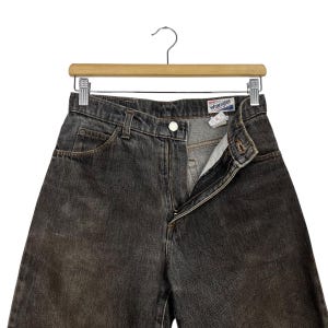 Puede incluir: Vaqueros Wrangler de mezclilla gris oscuro colgados de una percha de madera. Los vaqueros tienen cierre de bot&oacute;n y cremallera, con la cremallera parcialmente abierta. La etiqueta Wrangler es visible en la cintura. Los vaqueros est&aacute;n descoloridos y muestran signos de desgaste.