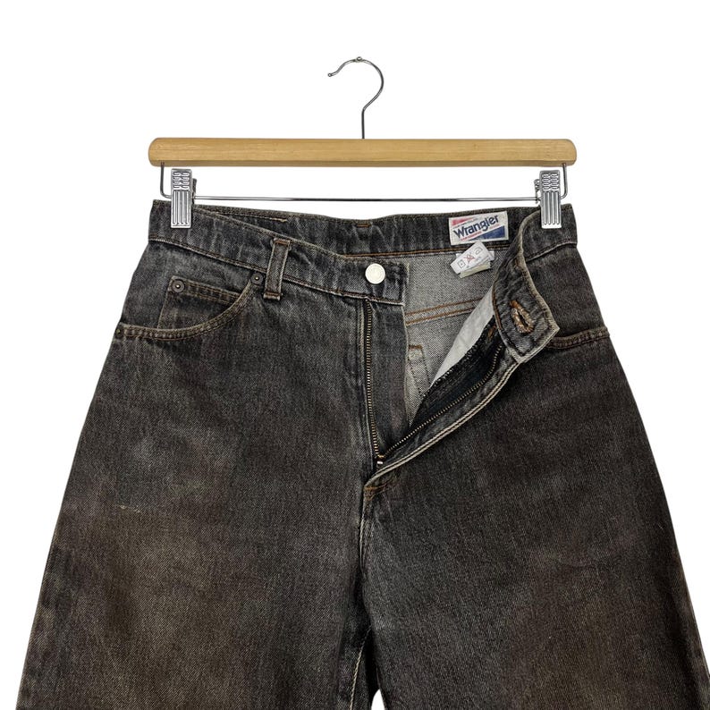Puede incluir: Vaqueros Wrangler de mezclilla gris oscuro colgados de una percha de madera. Los vaqueros tienen cierre de bot&oacute;n y cremallera, con la cremallera parcialmente abierta. La etiqueta Wrangler es visible en la cintura. Los vaqueros est&aacute;n descoloridos y muestran signos de desgaste.