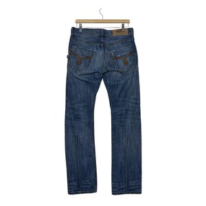 K&ouml;nnte beinhalten: Blaue Jeans aus Denim mit braunen N&auml;hten und Used-Details. Die Jeans haben ein klassisches F&uuml;nf-Taschen-Design, ein gerades Bein und eine geteilte Naht am unteren Ende der Beine. Die Jeans h&auml;ngen an einem Holzb&uuml;gel.