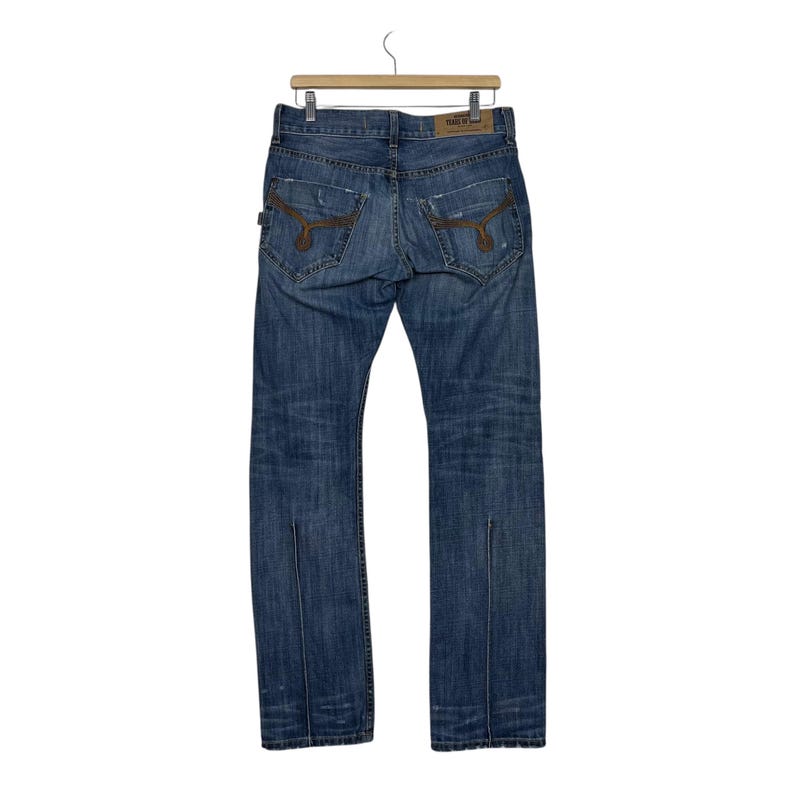 K&ouml;nnte beinhalten: Blaue Jeans aus Denim mit braunen N&auml;hten und Used-Details. Die Jeans haben ein klassisches F&uuml;nf-Taschen-Design, ein gerades Bein und eine geteilte Naht am unteren Ende der Beine. Die Jeans h&auml;ngen an einem Holzb&uuml;gel.