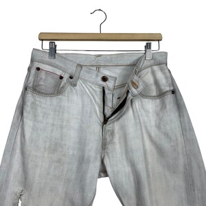 May include: Light gray denim jeans with a button and zipper closure, hanging on a wooden hanger. The jeans have a classic five-pocket design with a red accent on one pocket. The denim has a faded, worn look.