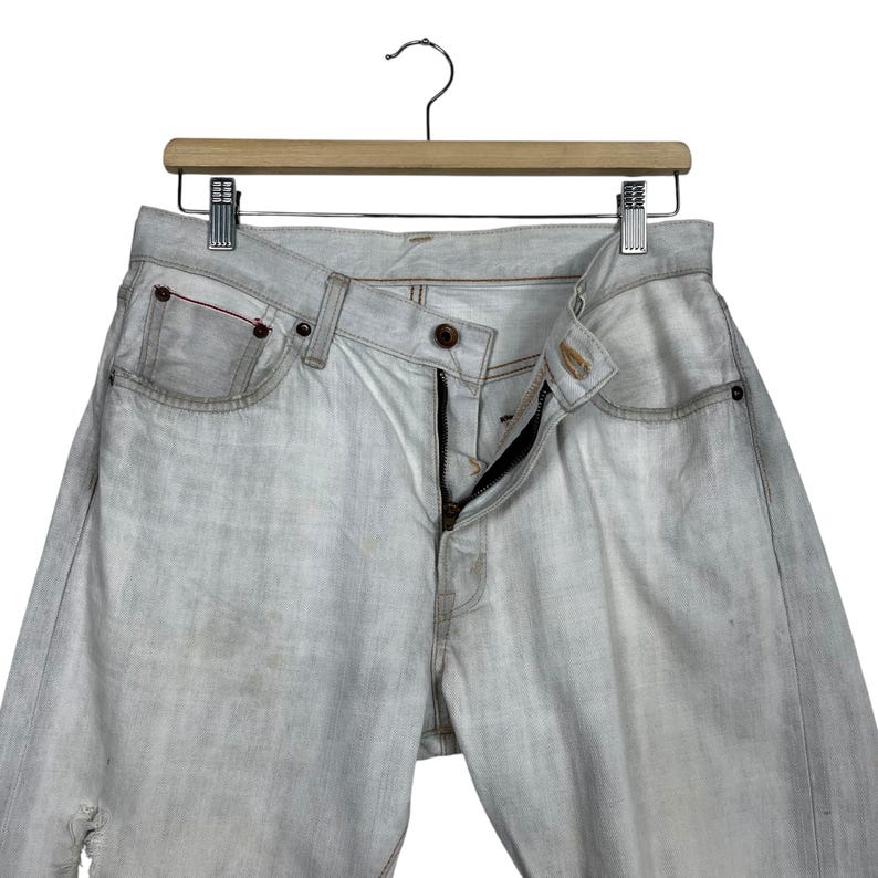 May include: Light gray denim jeans with a button and zipper closure, hanging on a wooden hanger. The jeans have a classic five-pocket design with a red accent on one pocket. The denim has a faded, worn look.