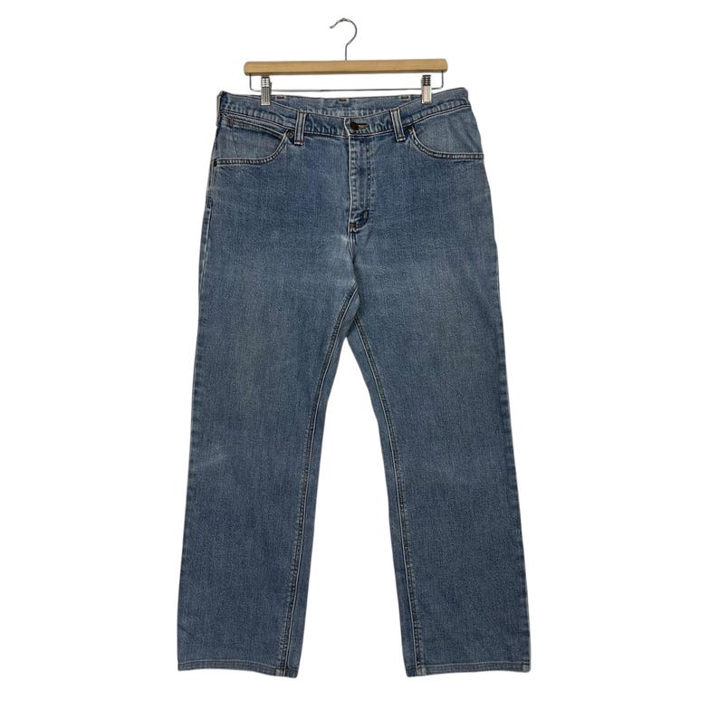 K&ouml;nnte beinhalten: Hellblaue Jeans aus Denim, die an einem Holzb&uuml;gel h&auml;ngen. Die Jeans haben ein klassisches F&uuml;nf-Taschen-Design, einen Knopf- und Rei&szlig;verschluss und eine gerade Beinpassform. Die Jeans sind aus einem strapazierf&auml;higen Denim-Stoff gefertigt.