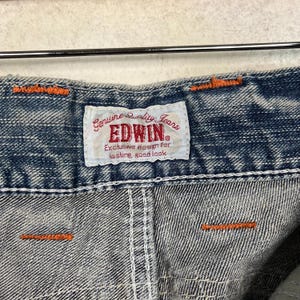 May include: Close-up of denim fabric with orange stitching and a white label. The label reads "Genuine Quality Jeans EDWIN Exclusive design for lasting good look."