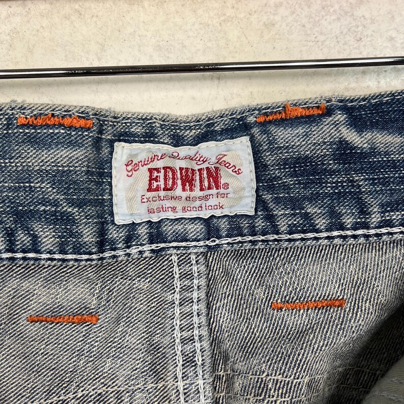 May include: Close-up of denim fabric with orange stitching and a white label. The label reads "Genuine Quality Jeans EDWIN Exclusive design for lasting good look."