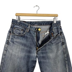 May include: Blue denim jeans with a button and zip fastening, hanging on a wooden hanger. The jeans have a classic five-pocket design and a faded wash. The button is gold-coloured.