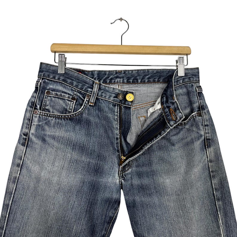 May include: Blue denim jeans with a button and zip fastening, hanging on a wooden hanger. The jeans have a classic five-pocket design and a faded wash. The button is gold-coloured.