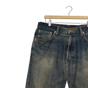 May include: Blue denim jeans with a faded wash, hanging on a wooden hanger. The jeans have classic five-pocket styling, a button and zipper closure, and visible stitching details. The denim has a worn, vintage appearance.