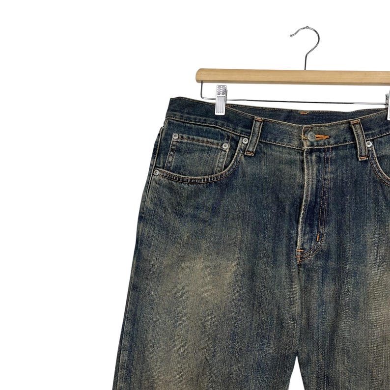 May include: Blue denim jeans with a faded wash, hanging on a wooden hanger. The jeans have classic five-pocket styling, a button and zipper closure, and visible stitching details. The denim has a worn, vintage appearance.
