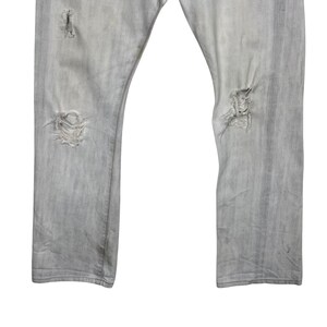 May include: Light gray denim jeans with distressed details. The jeans have multiple rips and tears, giving them a worn and vintage look. The fabric appears faded, adding to the overall aesthetic.