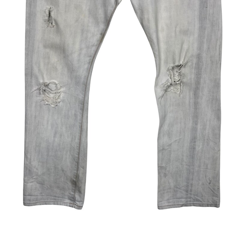 May include: Light gray denim jeans with distressed details. The jeans have multiple rips and tears, giving them a worn and vintage look. The fabric appears faded, adding to the overall aesthetic.