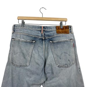 May include: Light blue denim jeans with distressed details, hanging on a wooden hanger. The jeans feature a leather patch that reads "EDWIN MADE IN JAPAN" and a red tag. The back pockets have a distinctive stitching design.