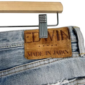May include: Close-up of light blue denim jeans with a brown leather patch. The patch features the brand name "EDWIN" and the text "MADE IN JAPAN". The jeans are hanging on a wooden hanger with a metal clip.