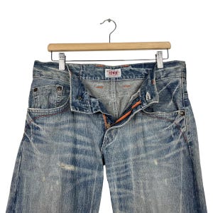May include: Light blue denim jeans with a distressed wash, featuring orange stitching and a visible zipper. The jeans are displayed on a wooden hanger, showcasing the front of the garment. The brand name "EDWIN" is visible on the waistband.