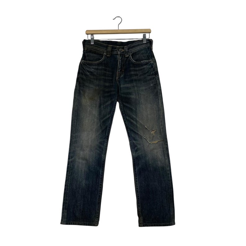 May include: Dark wash denim jeans with a distressed look, hanging on a wooden hanger. The jeans have a faded appearance with a rip on the right knee. The jeans are a classic five-pocket style.