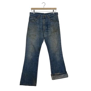 K&ouml;nnte beinhalten: Verblasste blaue Jeans mit Knopfleiste und ausgestelltem Bein. Die Jeans h&auml;ngen an einem Holzb&uuml;gel vor wei&szlig;em Hintergrund. Die Manschetten sind hochgekrempelt und zeigen einen helleren Denim-Farbton.