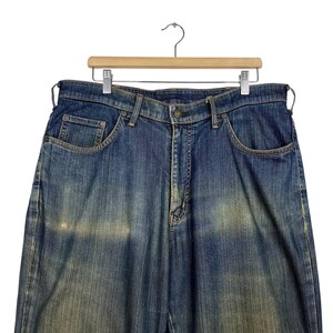 K&ouml;nnte beinhalten: Blaue Jeans aus Denim, die an einem Holzb&uuml;gel h&auml;ngen. Die Jeans haben ein klassisches F&uuml;nf-Taschen-Design, einen Knopf- und Rei&szlig;verschluss und eine verwaschene Optik. Die Jeans haben eine mittlere Waschung mit helleren Bereichen.