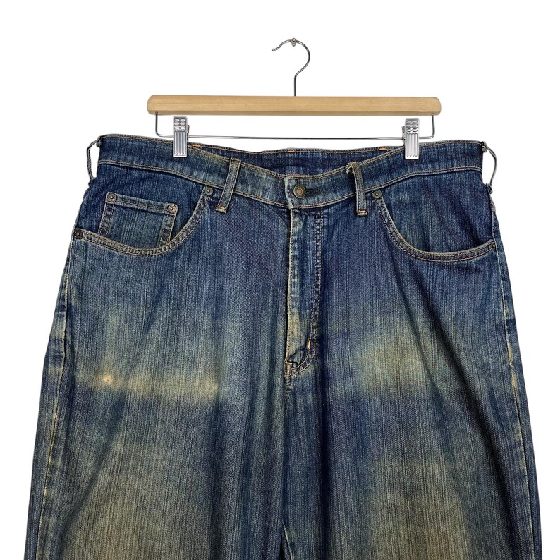 K&ouml;nnte beinhalten: Blaue Jeans aus Denim, die an einem Holzb&uuml;gel h&auml;ngen. Die Jeans haben ein klassisches F&uuml;nf-Taschen-Design, einen Knopf- und Rei&szlig;verschluss und eine verwaschene Optik. Die Jeans haben eine mittlere Waschung mit helleren Bereichen.