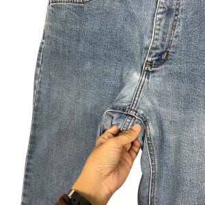 K&ouml;nnte beinhalten: Nahaufnahme einer hellblauen Jeans. Die Jeans hat ein klassisches F&uuml;nf-Taschen-Design mit Knopf- und Rei&szlig;verschluss. Die N&auml;hte sind sichtbar und der Stoff wirkt abgenutzt. Eine Hand h&auml;lt die Jeans.