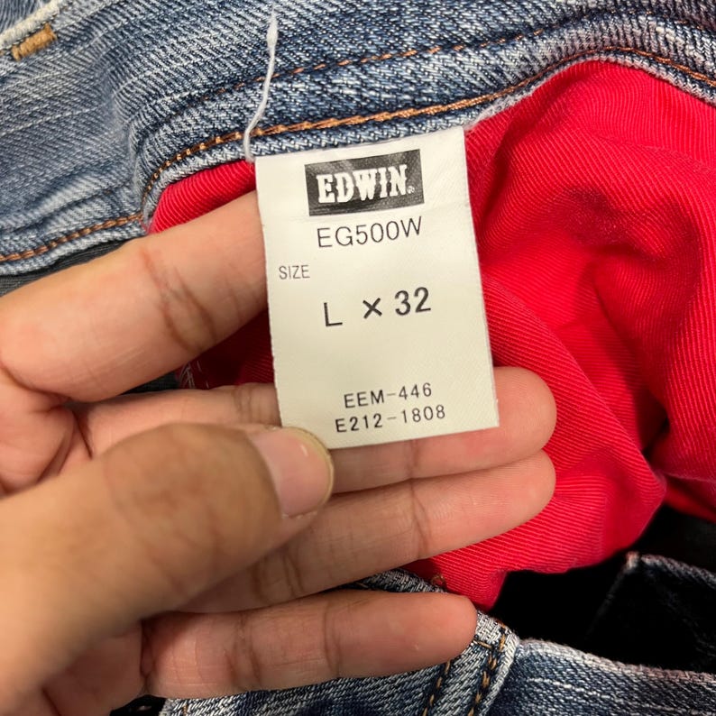 K&ouml;nnte beinhalten: Nahaufnahme eines Jeans-Etiketts mit dem Markennamen "EDWIN" in Wei&szlig; auf einem schwarzen Rechteck. Das Etikett zeigt auch "EG500W", die Gr&ouml;&szlig;e "L x 32" und andere Produktcodes. Die Jeans hat ein rotes Innenfutter.