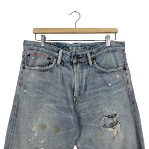 May include: Light blue denim jeans with distressed details, including rips and faded patches. The jeans are hanging on a wooden hanger with a metal clip. The jeans have a classic five-pocket design.