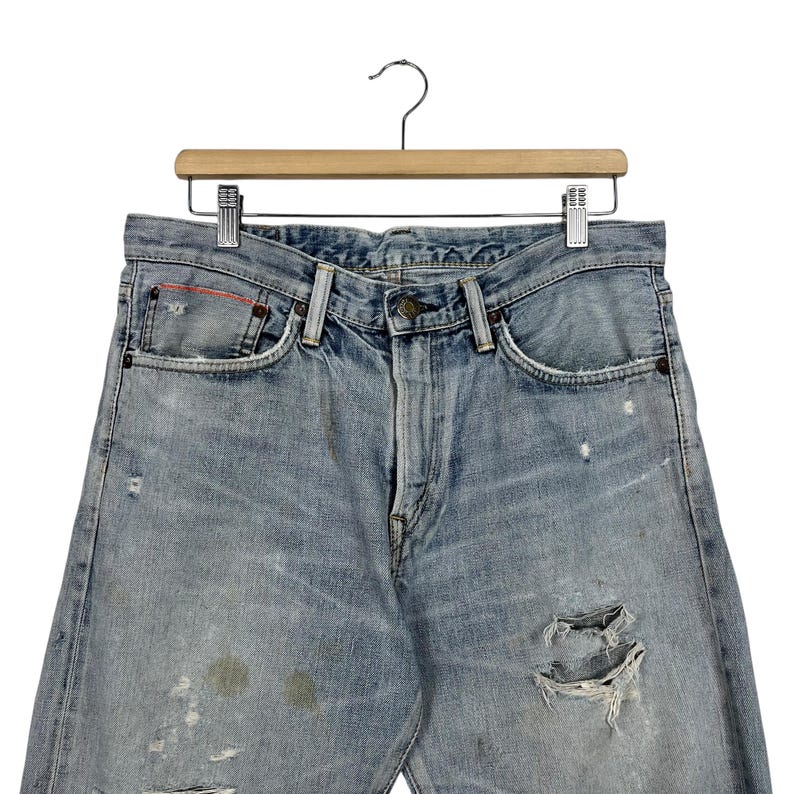 May include: Light blue denim jeans with distressed details, including rips and faded patches. The jeans are hanging on a wooden hanger with a metal clip. The jeans have a classic five-pocket design.