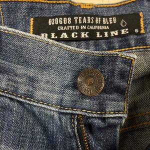 K&ouml;nnte beinhalten: Nahaufnahme einer blauen Jeans. Ein schwarzes Etikett mit der Aufschrift "020608 TEARS OF BLEU CRAFTED IN CALIFORNIA BLACK LINE". Ein kupferfarbener Knopf mit "TEARS OF BLEU" und der Nummer "020608" ist sichtbar.