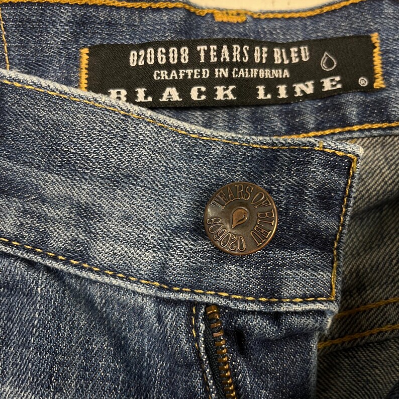 K&ouml;nnte beinhalten: Nahaufnahme einer blauen Jeans. Ein schwarzes Etikett mit der Aufschrift "020608 TEARS OF BLEU CRAFTED IN CALIFORNIA BLACK LINE". Ein kupferfarbener Knopf mit "TEARS OF BLEU" und der Nummer "020608" ist sichtbar.