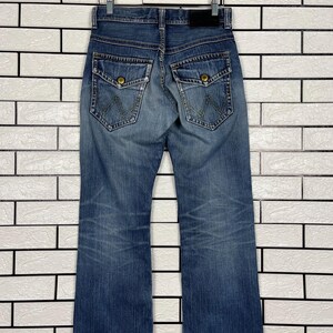 May include: Blue denim jeans with a classic five-pocket design, featuring a button and zipper closure. The jeans have a straight leg cut and are hanging on a wooden hanger against a white brick wall.