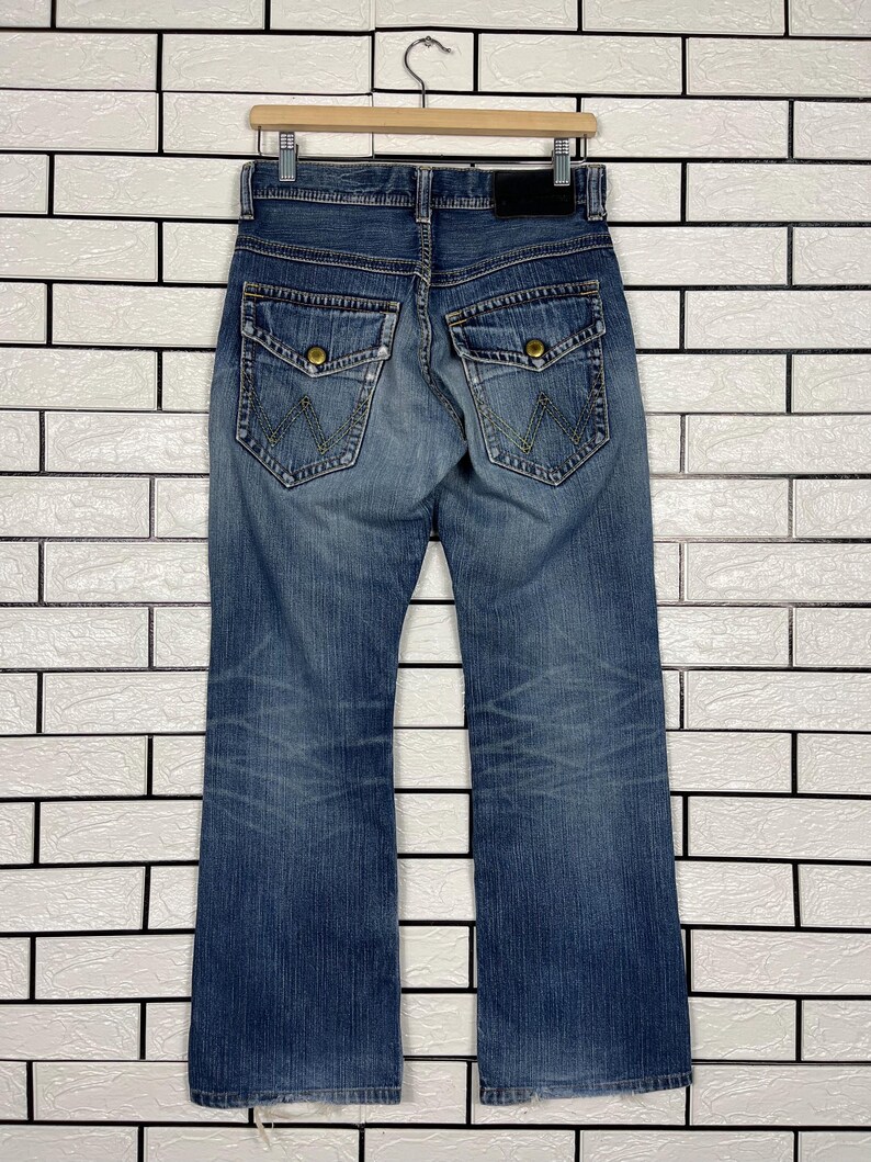 May include: Blue denim jeans with a classic five-pocket design, featuring a button and zipper closure. The jeans have a straight leg cut and are hanging on a wooden hanger against a white brick wall.
