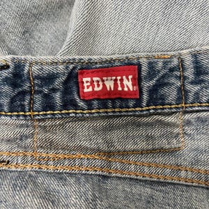 May include: Close-up of denim fabric with a red Edwin label. The label is rectangular with white text. The denim has visible stitching in a contrasting yellow-brown thread.