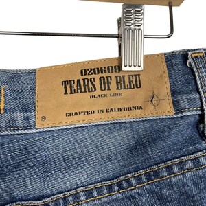K&ouml;nnte beinhalten: Nahaufnahme einer blauen Jeans mit einem braunen Lederetikett. Auf dem Etikett steht "TEARS OF BLEU BLACK LINE CRAFTED IN CALIFORNIA" in schwarzer Schrift. Die Jeans h&auml;ngt an einer Metallklammer.
