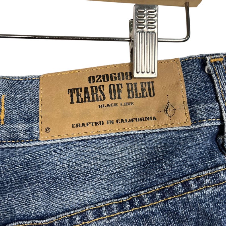 K&ouml;nnte beinhalten: Nahaufnahme einer blauen Jeans mit einem braunen Lederetikett. Auf dem Etikett steht "TEARS OF BLEU BLACK LINE CRAFTED IN CALIFORNIA" in schwarzer Schrift. Die Jeans h&auml;ngt an einer Metallklammer.