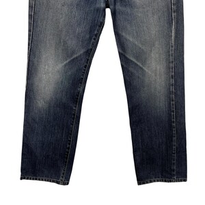 May include: Dark blue denim jeans with a straight leg cut. The jeans feature a classic five-pocket design and a button and zip closure. The denim has a worn appearance with lighter areas on the thighs and knees.
