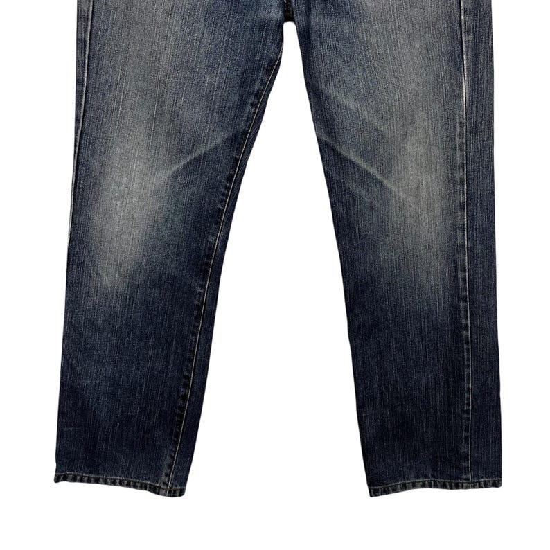 May include: Dark blue denim jeans with a straight leg cut. The jeans feature a classic five-pocket design and a button and zip closure. The denim has a worn appearance with lighter areas on the thighs and knees.
