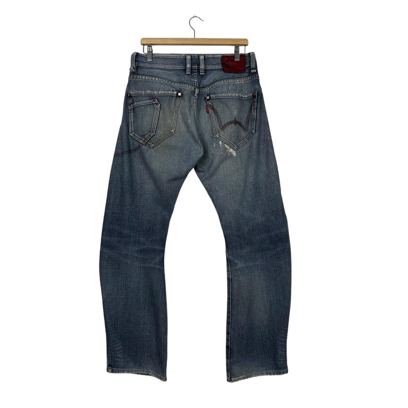 K&ouml;nnte beinhalten: Verblasste blaue Jeans mit einem Used-Look, h&auml;ngen an einem Holzb&uuml;gel. Die Jeans haben einen roten Lederaufn&auml;her und einzigartige Taschenmuster. Die Ges&auml;&szlig;taschen haben eine markante eckige Form.