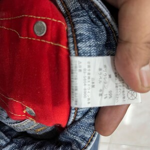 Op de afbeelding: Close-up van een blauwe spijkerbroek met een rode binnenvoering. Een wit label met Japanse tekst is zichtbaar, samen met een zilveren knoop. De jeans is gemaakt van 99% katoen en 1% polyurethaan.