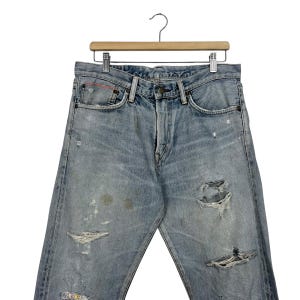May include: Light blue denim jeans with distressed details, including rips and faded patches. The jeans are hanging on a wooden hanger. The jeans have a classic five-pocket design and a button and zipper closure.