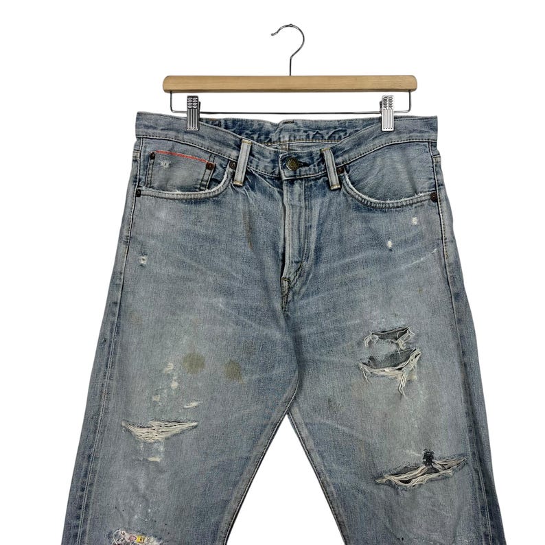 May include: Light blue denim jeans with distressed details, including rips and faded patches. The jeans are hanging on a wooden hanger. The jeans have a classic five-pocket design and a button and zipper closure.