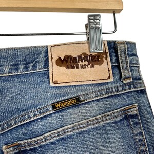 K&ouml;nnte beinhalten: Nahaufnahme einer hellblauen Jeans mit dem Wrangler-Logo auf der Ges&auml;&szlig;tasche und einem Lederaufn&auml;her mit der Aufschrift "Wrangler AMERICA". Die Jeans h&auml;ngt an einer Metallklammer an einem Holzb&uuml;gel.