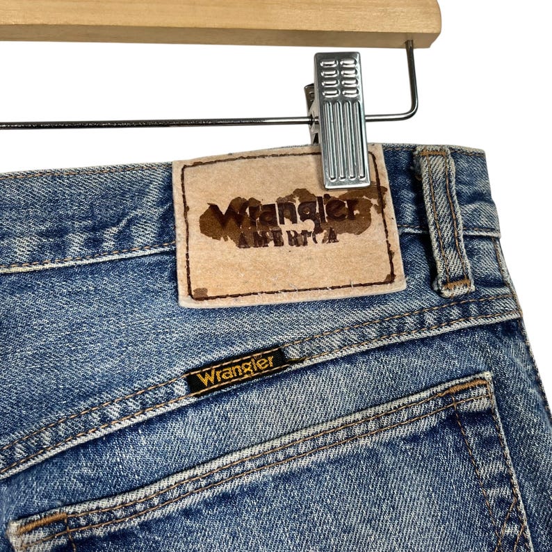 K&ouml;nnte beinhalten: Nahaufnahme einer hellblauen Jeans mit dem Wrangler-Logo auf der Ges&auml;&szlig;tasche und einem Lederaufn&auml;her mit der Aufschrift "Wrangler AMERICA". Die Jeans h&auml;ngt an einer Metallklammer an einem Holzb&uuml;gel.