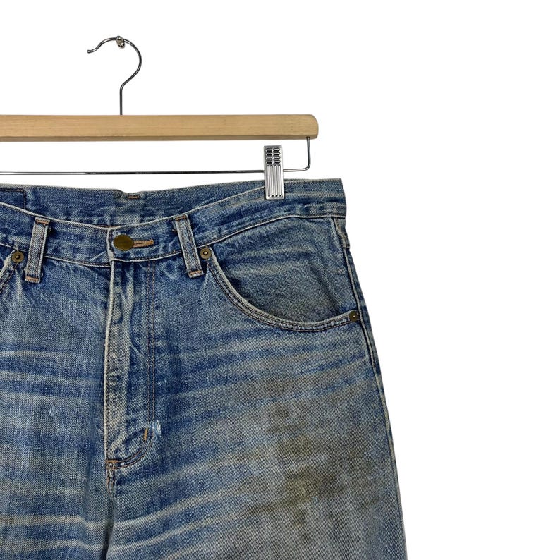 K&ouml;nnte beinhalten: Nahaufnahme einer verblichenen blauen Jeans, die an einem Holzb&uuml;gel h&auml;ngt. Die Jeans weist Gebrauchsspuren mit leichten Abnutzungen und Verf&auml;rbungen auf. Die Jeans hat ein klassisches F&uuml;nf-Taschen-Design.