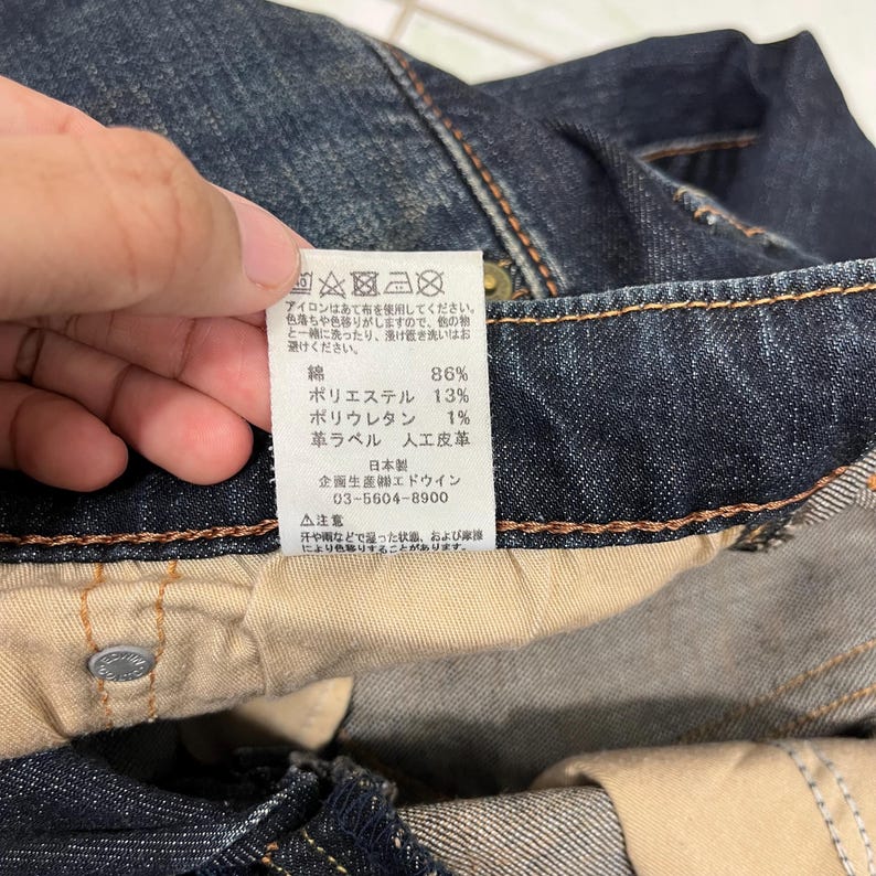 Puede incluir: Primer plano de unos vaqueros de mezclilla de lavado oscuro. Se ve una etiqueta blanca con texto japon&eacute;s, que detalla la composici&oacute;n de la tela y las instrucciones de cuidado. Los vaqueros tienen un dise&ntilde;o cl&aacute;sico de cinco bolsillos y cierre de bot&oacute;n.