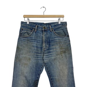 K&ouml;nnte beinhalten: Verblasste blaue Jeans aus Denim, die an einem Holzb&uuml;gel h&auml;ngen. Die Jeans haben ein klassisches F&uuml;nf-Taschen-Design, einen Knopf- und Rei&szlig;verschluss und weisen Gebrauchsspuren mit Verf&auml;rbungen auf.