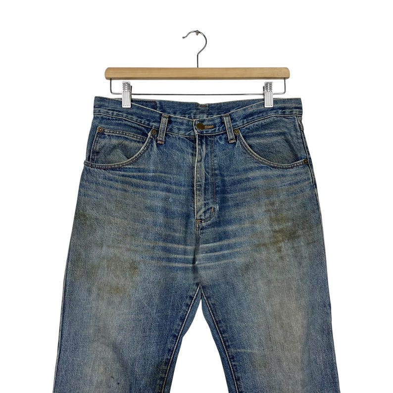 K&ouml;nnte beinhalten: Verblasste blaue Jeans aus Denim, die an einem Holzb&uuml;gel h&auml;ngen. Die Jeans haben ein klassisches F&uuml;nf-Taschen-Design, einen Knopf- und Rei&szlig;verschluss und weisen Gebrauchsspuren mit Verf&auml;rbungen auf.